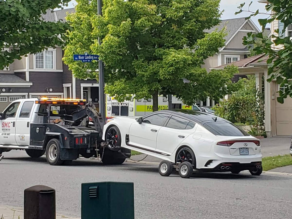 A white SMC tow truck towing a white sports sedan on a residential street, with an ambulance, River Landing Ave street sign, and suburban houses in the background under green trees.