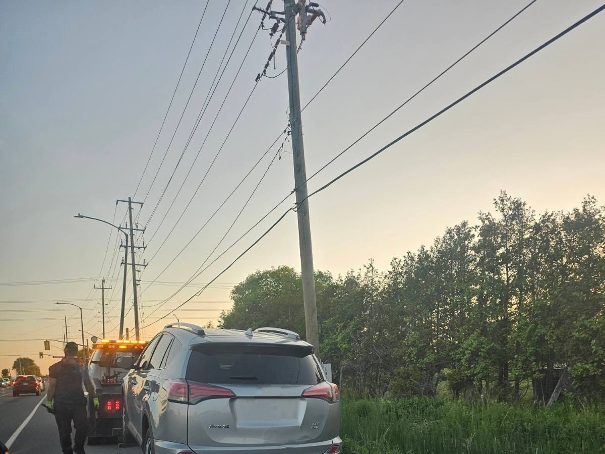 Dusk roadside scene with a silver SUV being towed by a tow truck with flashing warning lights, a tow worker stands near the vehicles, utility poles with overhead power lines line the road, with trees and other traffic visible in the background
