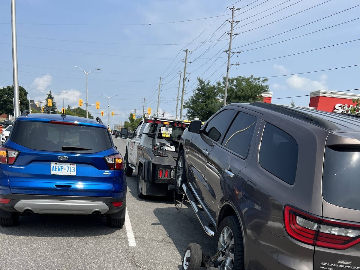 Daytime roadside scene at a signalized intersection, featuring a blue Ford Escape, a tow truck towing a dark grey Dodge Durango, with overhead power lines and a retail store sign in the background.
